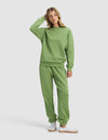 A woman in the Cozy Earth Women's Cityscape 2.0 Crewneck & Sweatpant Set, light green, stands casually with one hand touching her head. She pairs the outfit with white sneakers and looks slightly to the side against a plain background.|Color:Clover