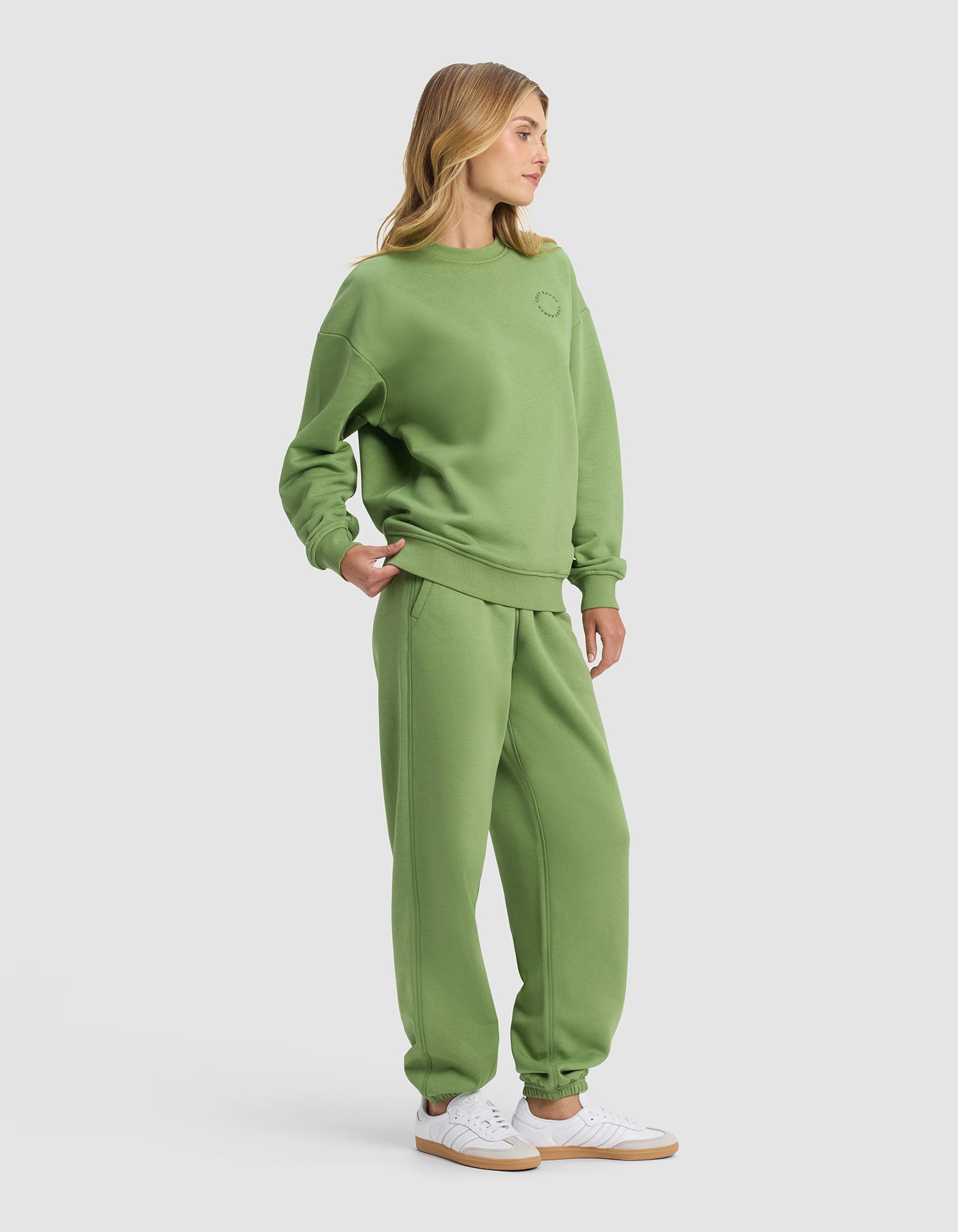 A woman models the Cozy Earth Women's Cityscape 2.0 Crewneck & Sweatpant Set in green with white sneakers against a plain light gray background, standing slightly to the side.