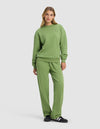 A woman poses casually in the Cozy Earth Women's Cityscape 2.0 Crewneck & Wide Leg Pant Set, featuring a relaxed fit. She stands against a plain background with one hand in her pocket, completing the look with black and white sneakers.|Color:Clover