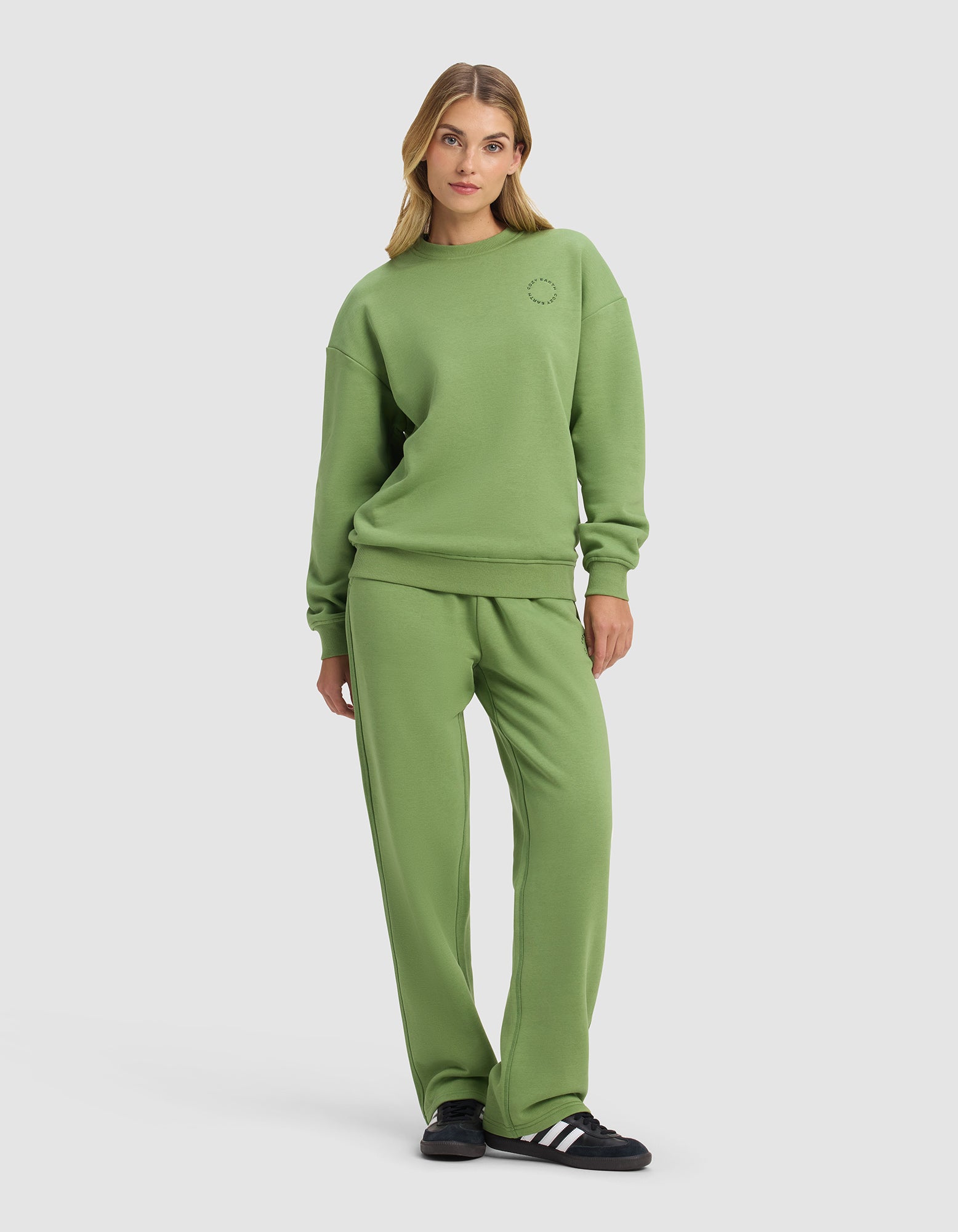 A woman poses casually in the Cozy Earth Women's Cityscape 2.0 Crewneck & Wide Leg Pant Set, featuring a relaxed fit. She stands against a plain background with one hand in her pocket, completing the look with black and white sneakers.