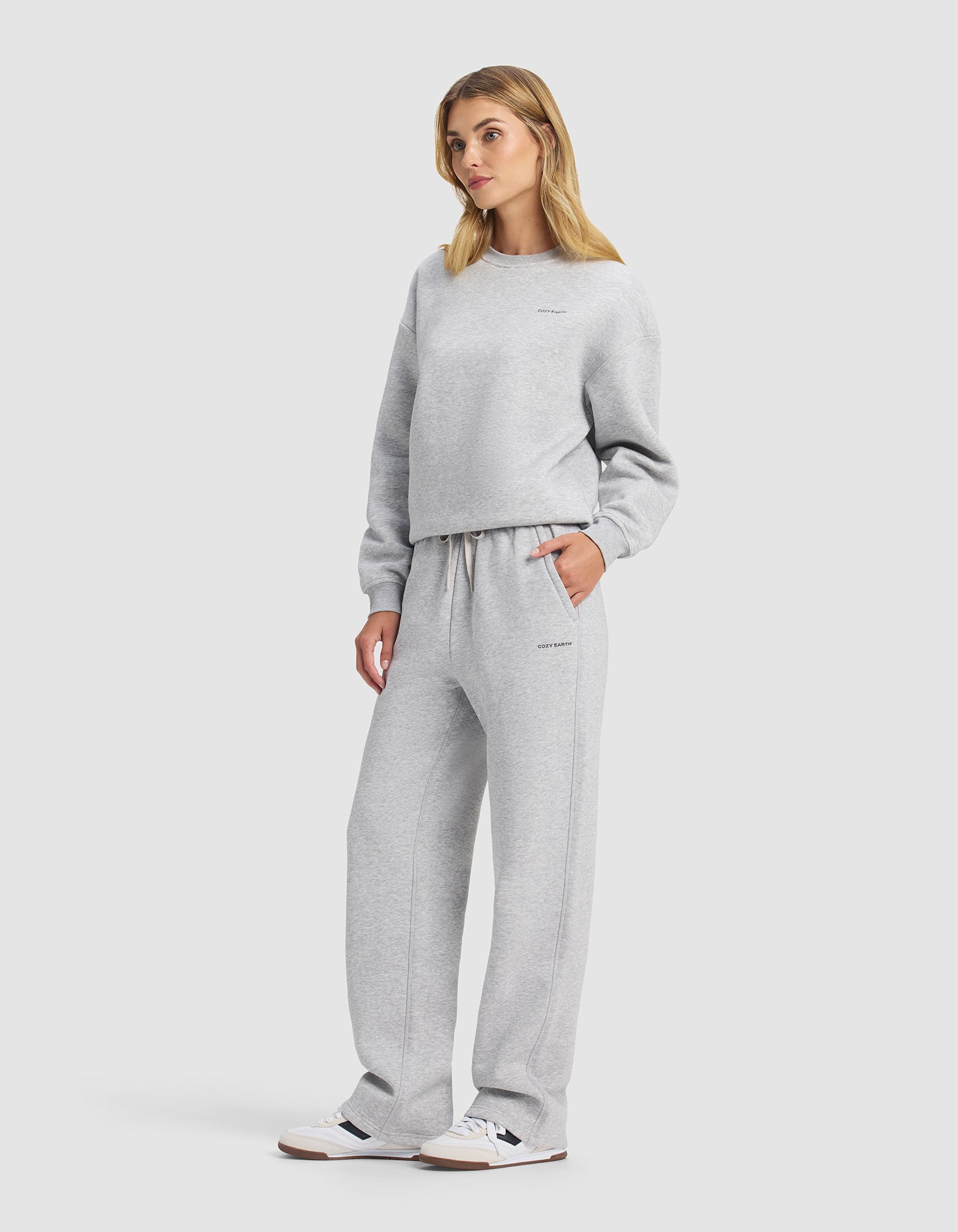 A woman wears the Cozy Earth Women's Cityscape 2.0 Crewneck & Wide Leg Pant Set in light gray with white sneakers, hands in pockets, blonde hair, standing against a plain white background.|Color:Heather Grey