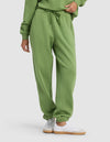 A person wears Cozy Earth's Women's Cityscape 2.0 Sweatpant—a green pair with an elastic waistband and drawstring—styled with a matching sweatshirt and white sneakers, standing against a plain light background.|Color:Clover