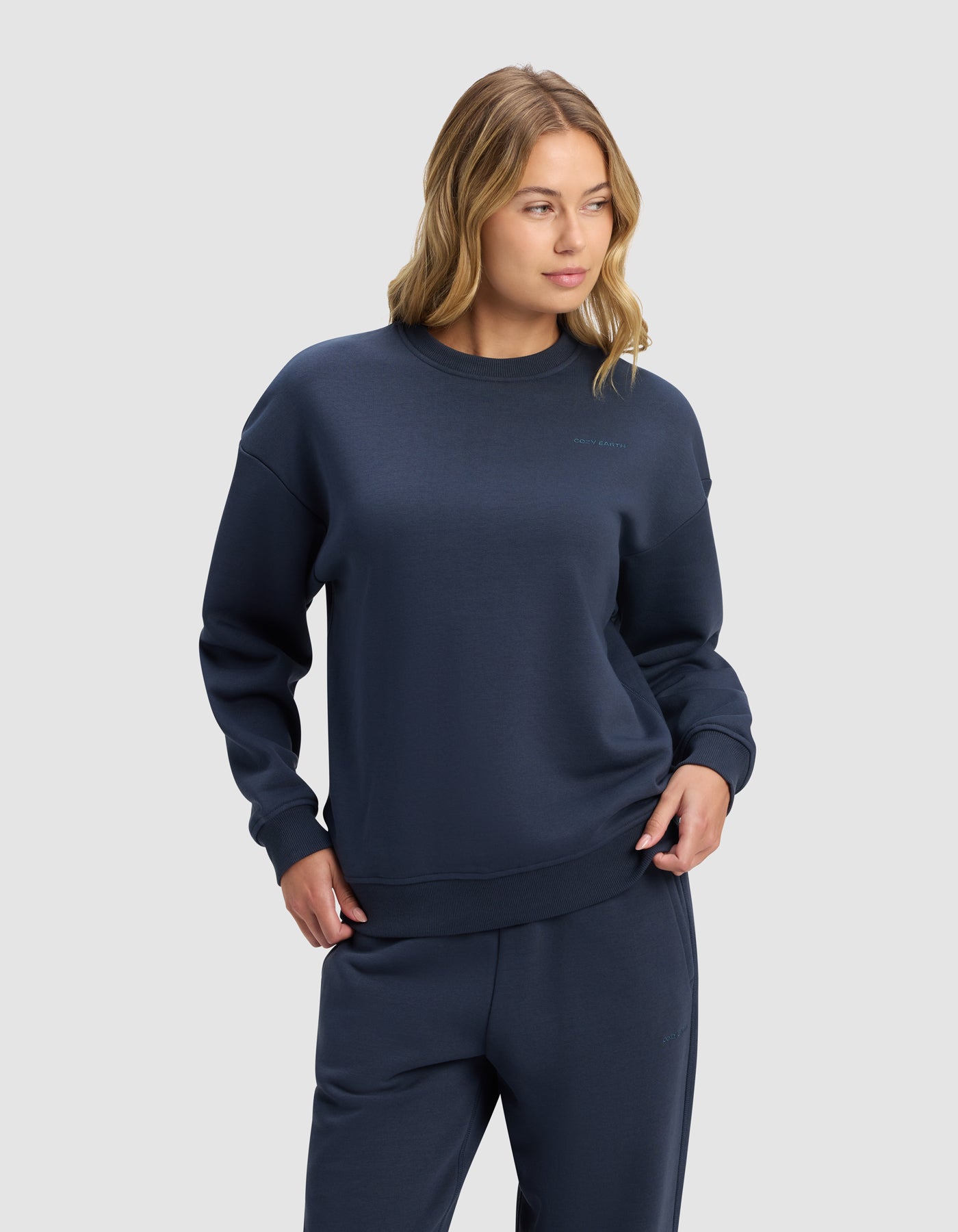 A woman with loose blonde waves wears the Cozy Earth Women's Cityscape 2.0 Crewneck in dark blue, paired with matching sweatpants, standing confidently against a light gray background.