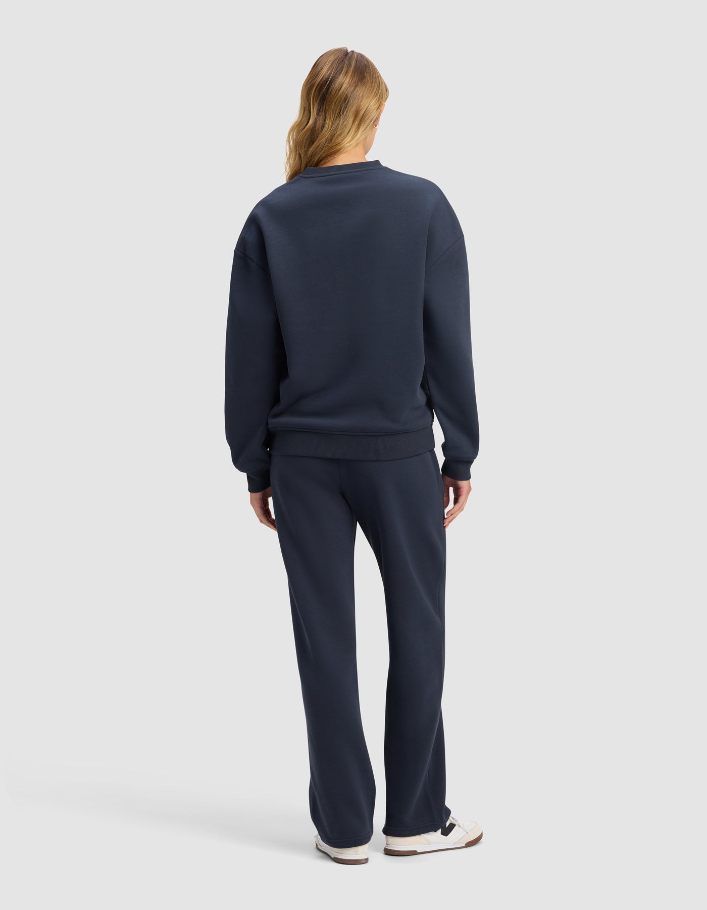 A woman with long, light brown hair stands facing away, wearing the Cozy Earth Women's Cityscape 2.0 Crewneck & Wide Leg Pant Set in navy blue with white sneakers against a plain light gray background.