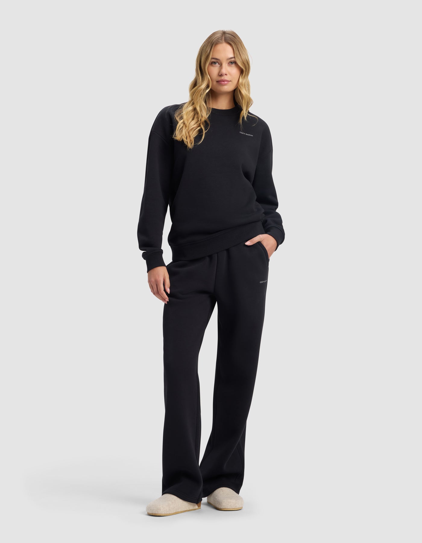 A woman with long blonde hair wears the Cozy Earth Women's Cityscape Crewneck & Wide Leg Pant Set in black, standing forward with one hand in her pocket and light-colored slippers, against a plain light background.|Color:Jet Black