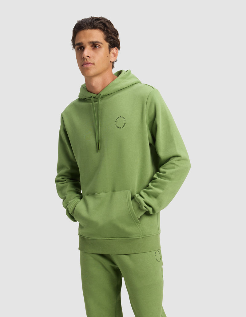 A man wears a Cozy Earth Men's Cityscape 2.0 Hoodie and matching sweatpants in Clover, both featuring a small circular stitched design, with hands in the hoodie pocket against a plain light gray background.