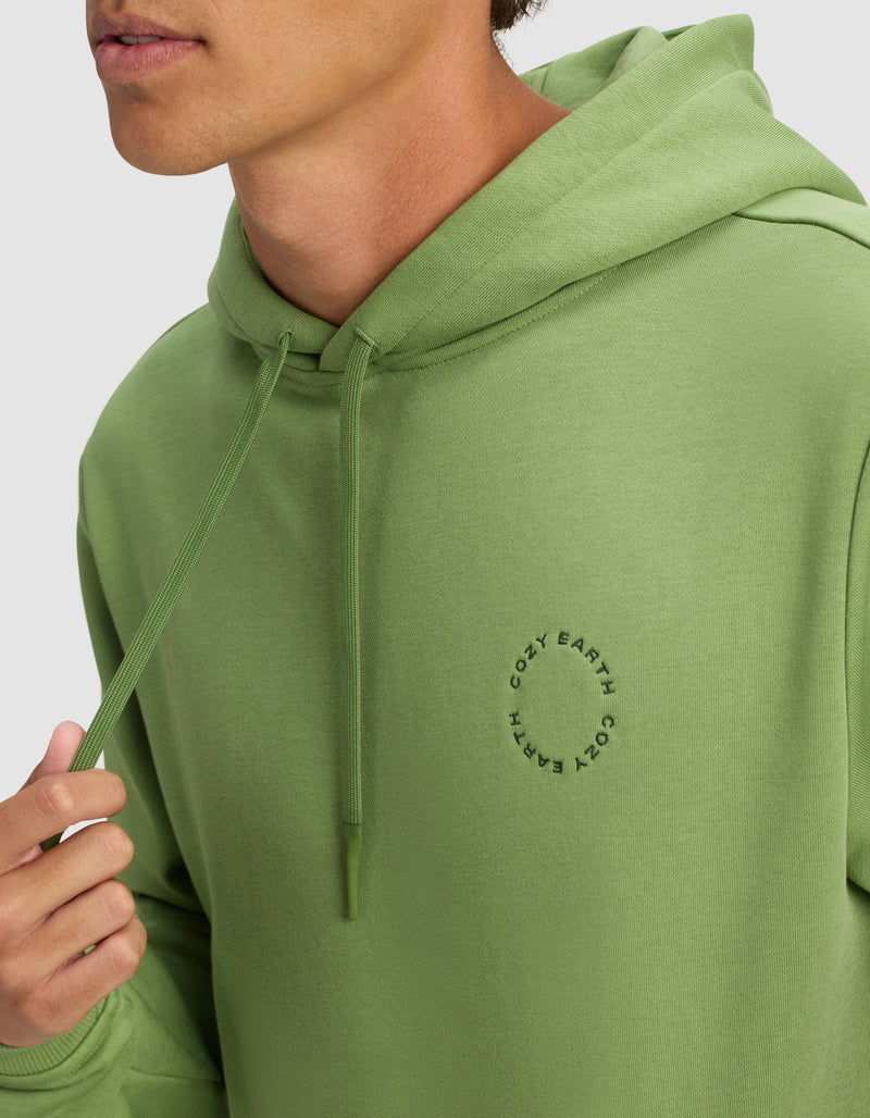 A man wears the Men's Cityscape Hoodie by Cozy Earth in light green, featuring a small circular logo on the chest. He holds a hoodie drawstring, with only his lower face visible.