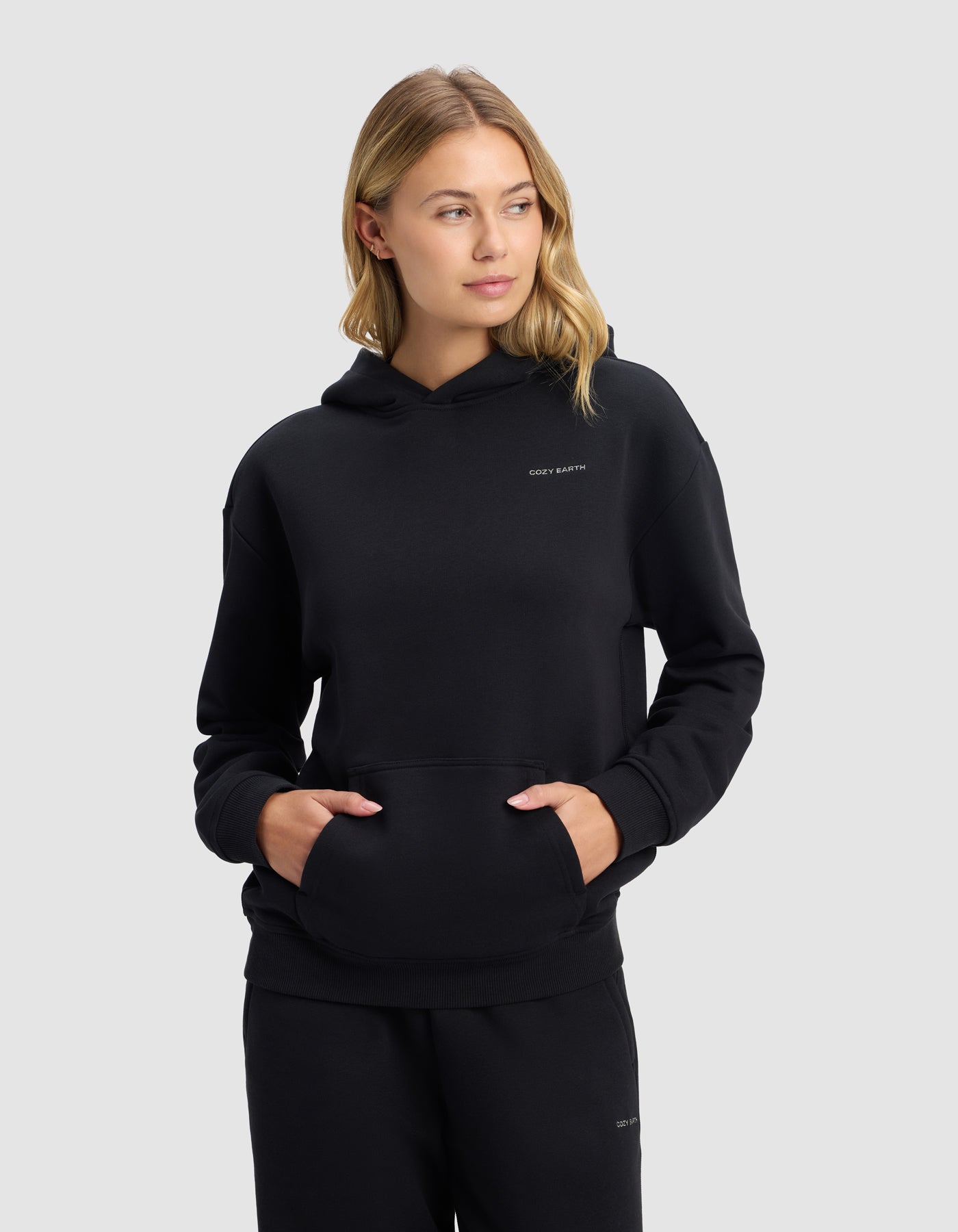 A young blonde woman models the Cozy Earth Women's Cityscape Hoodie in black with matching pants, standing with hands in her hoodie pocket and looking slightly left, against a plain light grey background.|Color:Jet Black