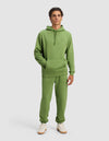 A young man wears the Cozy Earth Men's Cityscape 2.0 Hoodie & Sweatpant Set with white sneakers, standing against a plain background with his hands in the hoodie pocket and a neutral expression.|Color:Clover