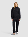A woman with long blonde hair models the Cozy Earth Women's Cityscape Hoodie & Sweatpant Set in black with grey slippers, standing against a plain light grey background.|Color:Jet Black