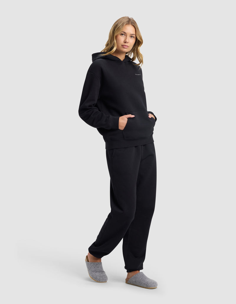 A woman with light brown hair poses against a light gray background wearing Cozy Earth's Women's Cityscape Hoodie & Sweatpant Set in black, with her hands in the front hoodie pocket and gray slippers on her feet.