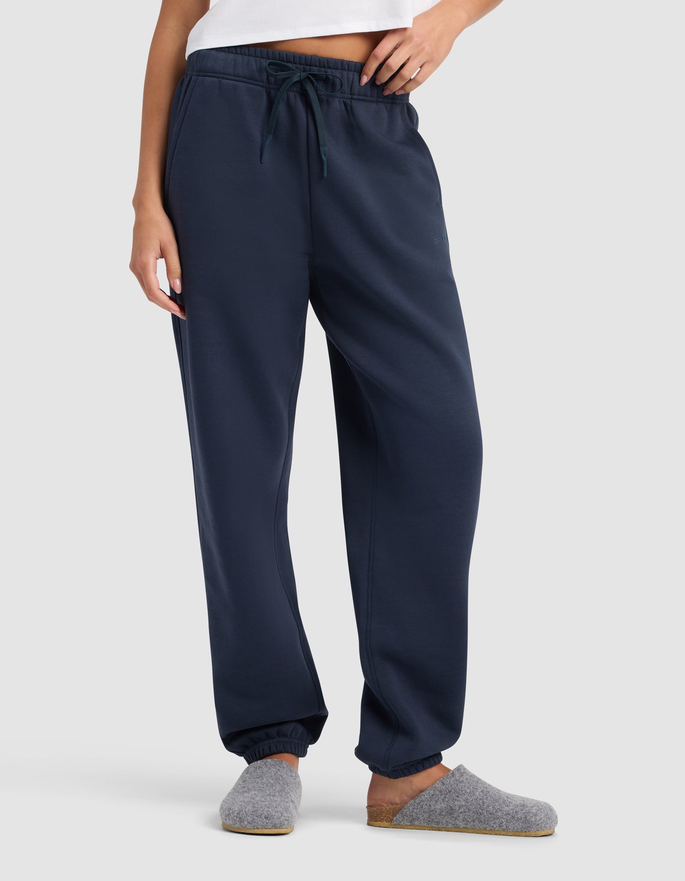 A woman stands against a plain light background wearing Cozy Earth’s Women’s Cityscape Sweatpant in navy blue with an elastic waistband and drawstring, paired with a white top and grey slippers.|Color:Eclipse