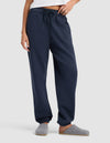A woman stands against a plain light background wearing Cozy Earth’s Women’s Cityscape Sweatpant in navy blue with an elastic waistband and drawstring, paired with a white top and grey slippers.|Color:Eclipse