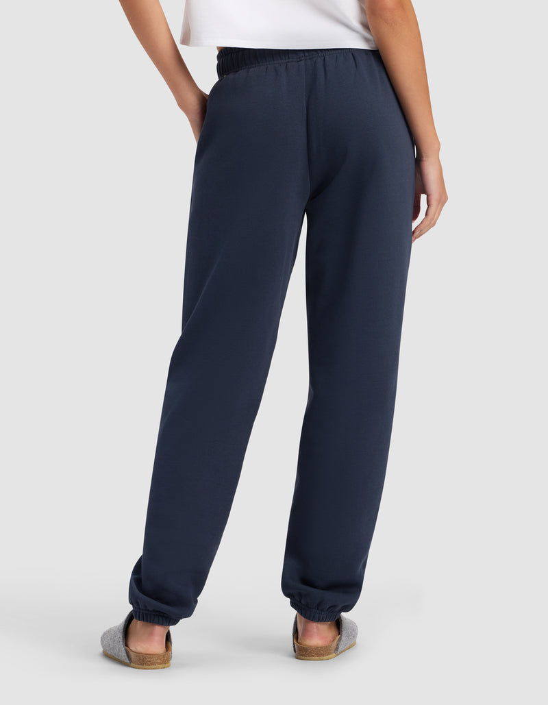 A person stands with their back to the camera, wearing a white shirt and Cozy Earth Women's Cityscape Sweatpant in navy blue, paired with gray slippers against a light background.