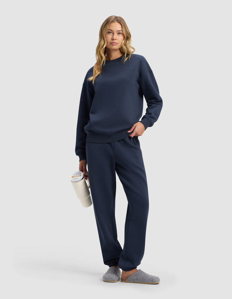 A woman in Cozy Earth's Women's Cityscape Sweatpant, paired with a navy blue sweatshirt and gray slippers, holds a white water bottle. She has long wavy blonde hair, a relaxed look, and stands against a plain light gray background.