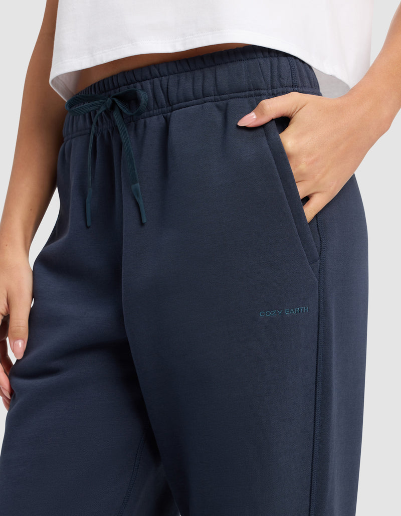 A person wears Cozy Earth Women's Cityscape Sweatpant in dark blue with one hand in the pocket, visible drawstrings, part of a white top shown, and "COZY EARTH" embroidered near the pocket.