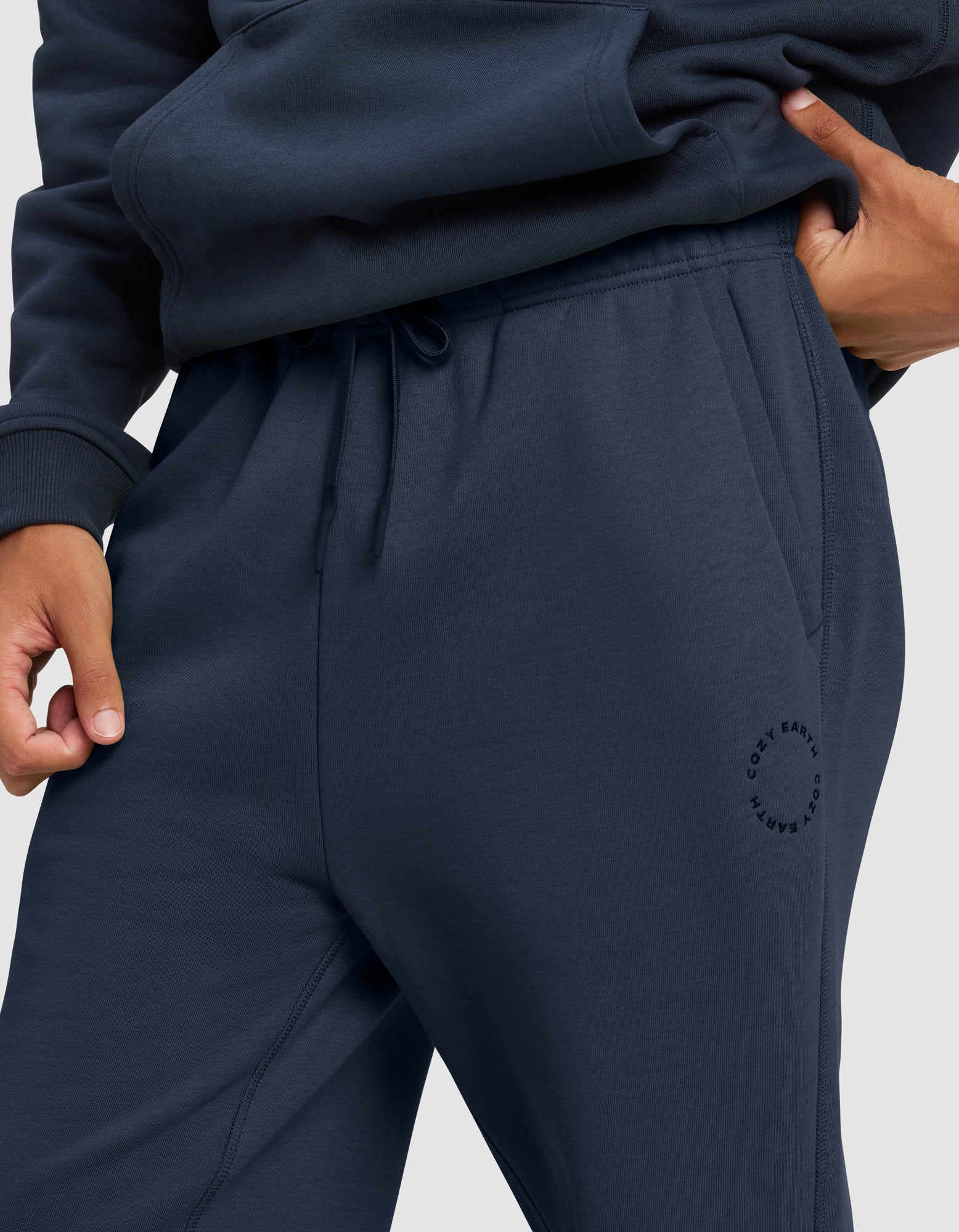 A person wears Cozy Earth Men's Cityscape 2.0 Sweatpants in navy blue with a drawstring and a circular "Cozy Earth" logo near the pocket, one hand tucked in, paired with a matching sweatshirt.