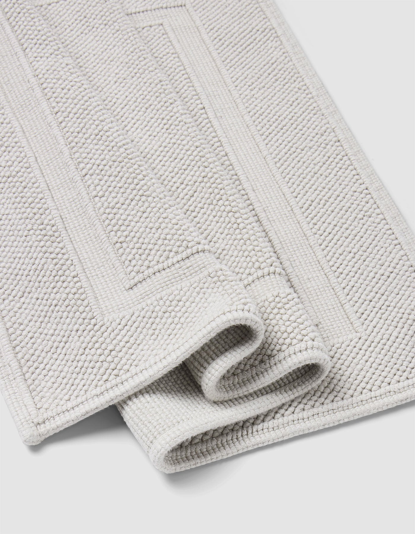 The Cozy Earth Classic Bath Mat is a folded light grey, textured mat with a rectangular border pattern, displayed on a white background.|Color:Light Grey