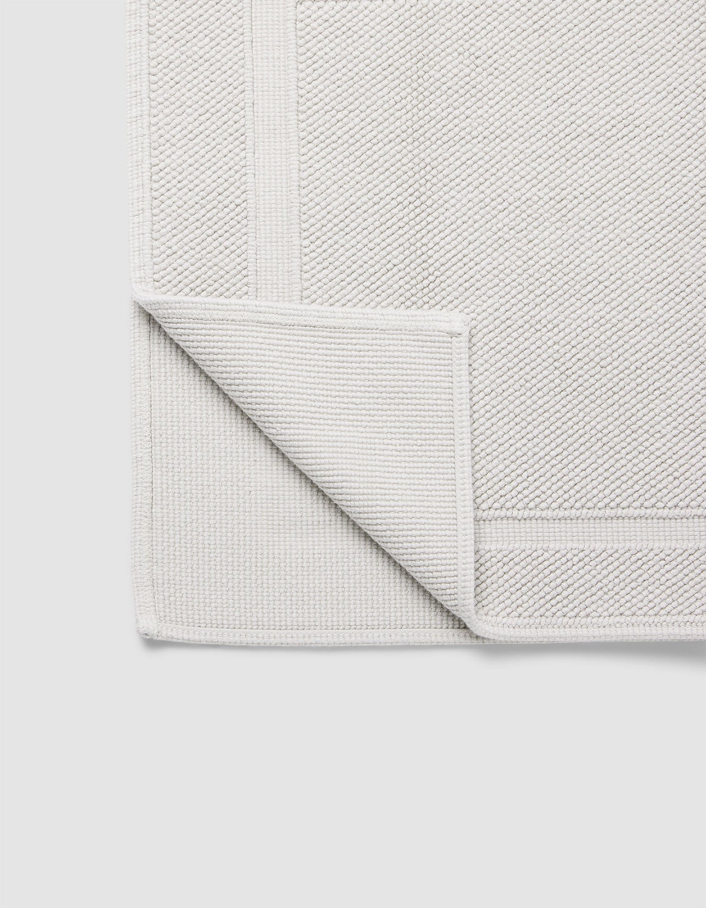 The Cozy Earth Classic Bath Mat in light grey is displayed flat on a plain white background, with one corner folded over to show its woven underside and detailed border.