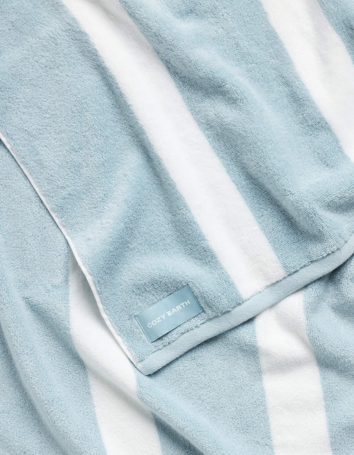 The Classic Stripe Resort Towel by Cozy Earth features plush fabric, neatly folded, with light blue and white vertical stripes and a sewn-in "COZY EARTH" tag on the edge.