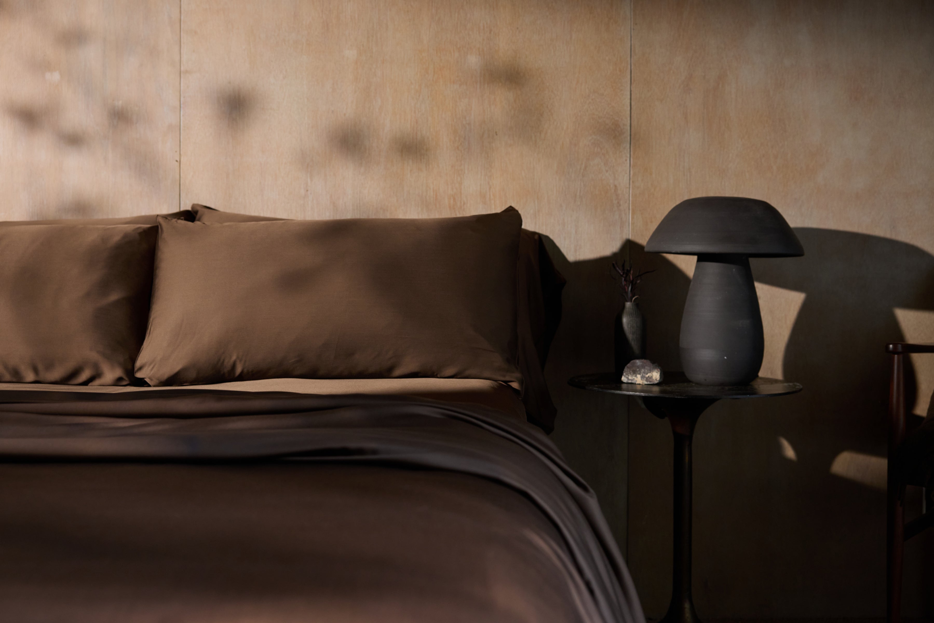 Brown satin bedding set on a bed with a matching nightstand lamp against a textured wall.