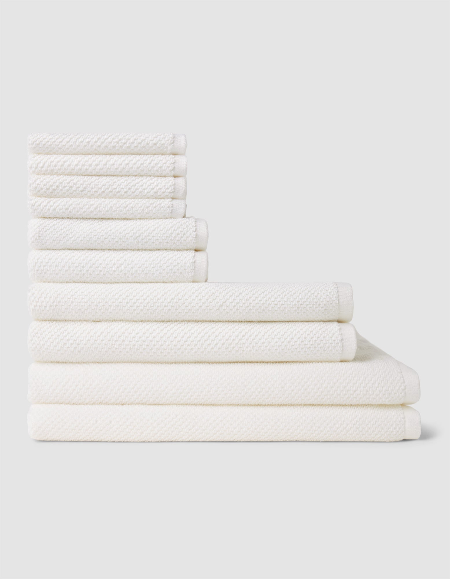 The Nantucket Bath Towel Set by Cozy Earth is shown neatly stacked by size from smallest to largest on a light gray background.|Color:White