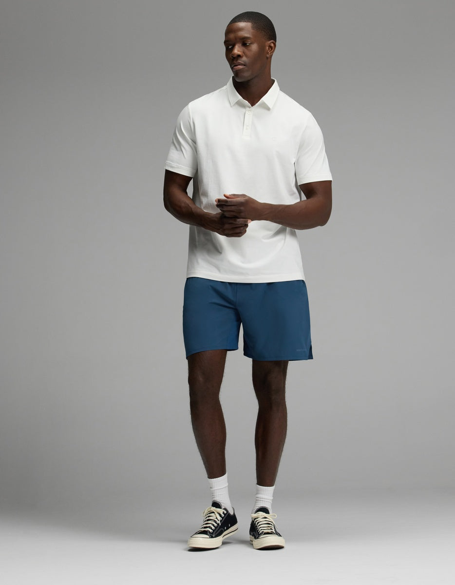 A man is standing in a studio wearing the Cozy Earth Men’s Day & Night Short 7″. He pairs them with a white polo shirt, white socks, and black sneakers. With his hands loosely clasped in front of him, he looks to the side against a plain gray background.