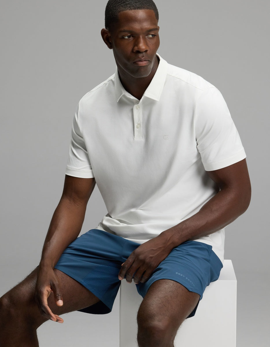 A man wearing Cozy Earth’s Men’s Day & Night Short 7″ in blue sits on a white block against a plain gray background, looking to his left with a neutral expression.