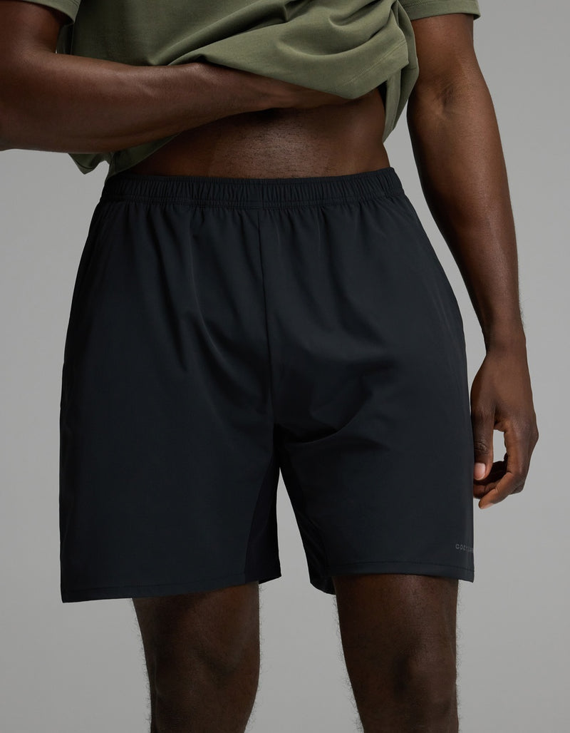 A person wearing a green shirt and the Cozy Earth Men’s Day & Night Short 7″ in Jet Black stands with one hand on their hip against a plain gray background.