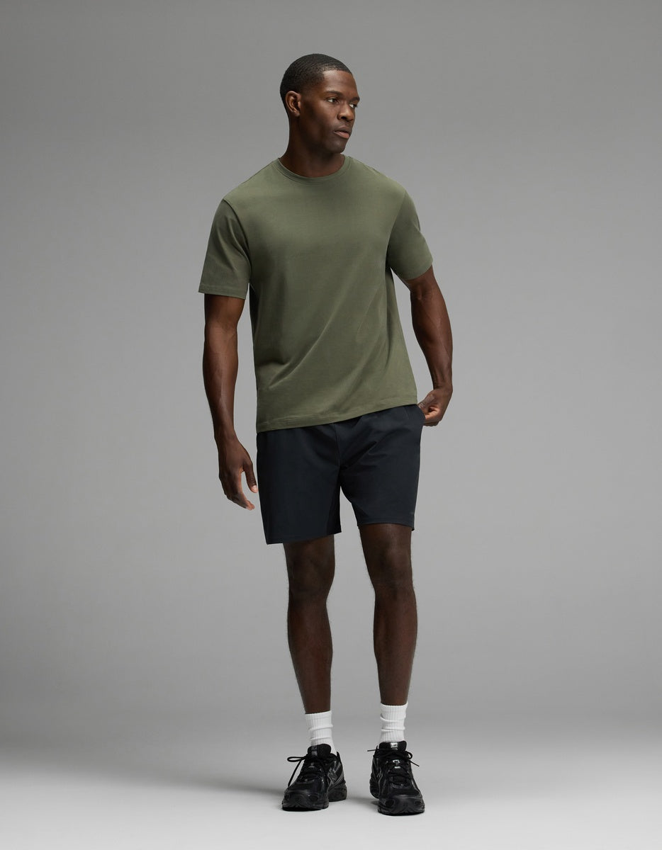 A man wearing Cozy Earth’s Men’s Day & Night Short 7″ in black, paired with an olive green t-shirt, white socks, and black sneakers stands against a plain gray background.