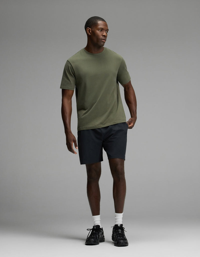 A man wearing Cozy Earth’s Men’s Day & Night Short 7″ in black, paired with an olive green t-shirt, white socks, and black sneakers stands against a plain gray background.