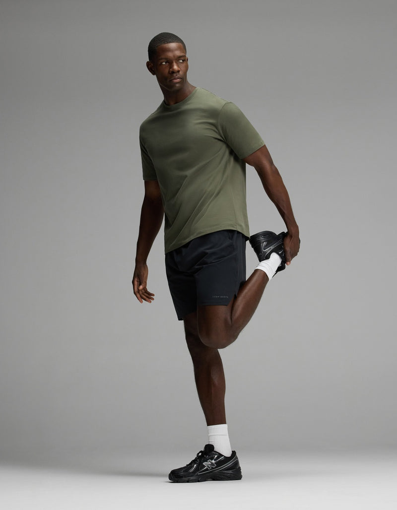 A man wearing Cozy Earth’s Men’s Day & Night Short 7″ in black and an olive green t-shirt stands on one leg, holding his ankle behind him in a quad stretch. He pairs the shorts with white socks and black sneakers, gazing off to the side against a gray background.