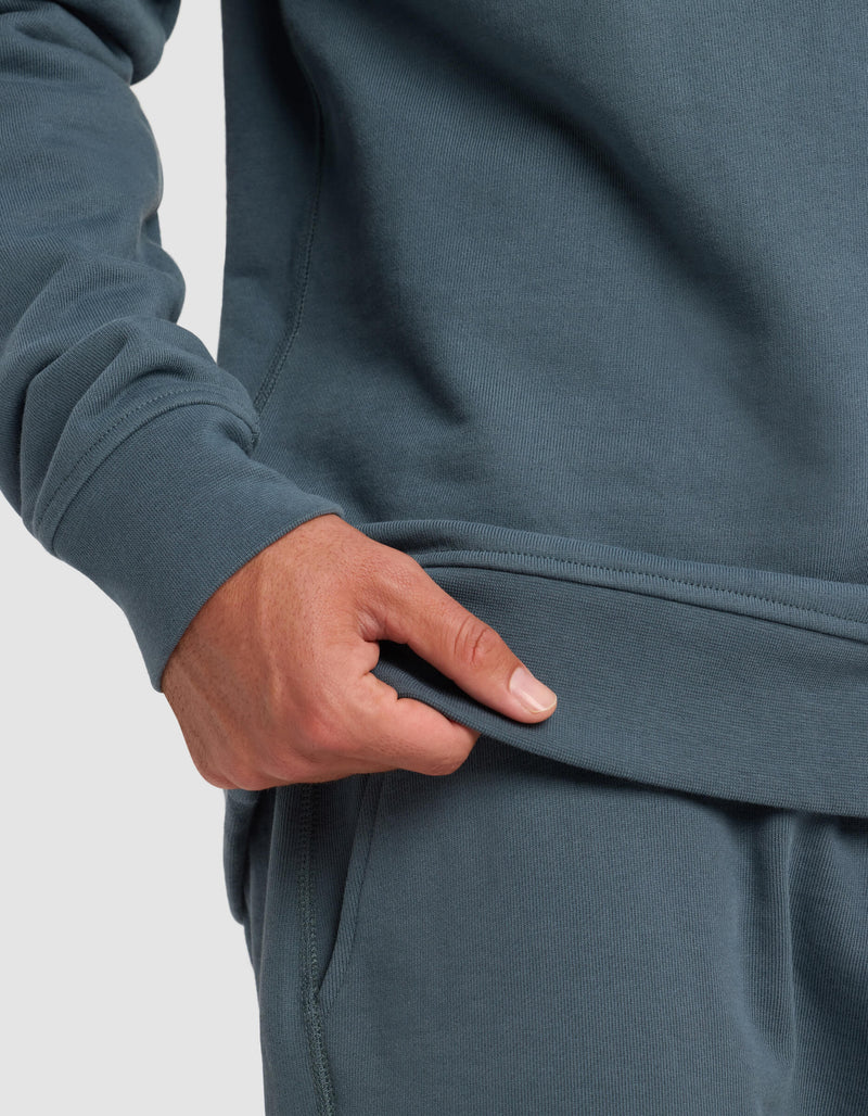 A person wearing the Cozy Earth Men's Dylan Crewneck in teal and matching sweatpants gently pulls at the sweatshirt hem with one hand.
