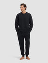 A man stands barefoot against a light gray background, wearing the Cozy Earth Men's Dylan Crewneck & Jogger Set in black. He has his hands in his pockets and gazes off to the side with a relaxed expression.|Color:Jet Black