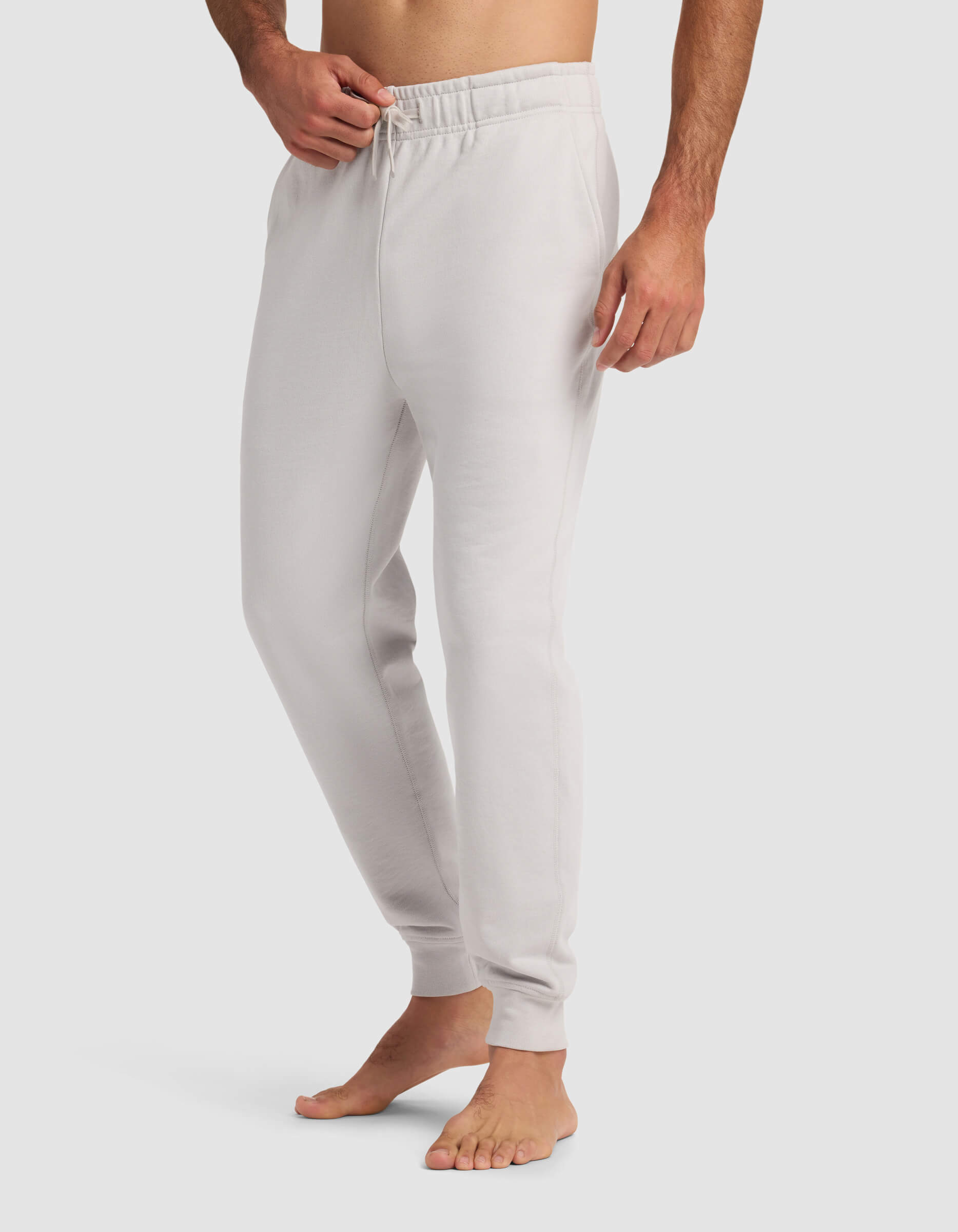 A barefoot man wears Cozy Earth's Men's Dylan Jogger in light gray, featuring an elastic waistband and drawstring, with one hand in his pocket. The background is plain white.|Color:Dew