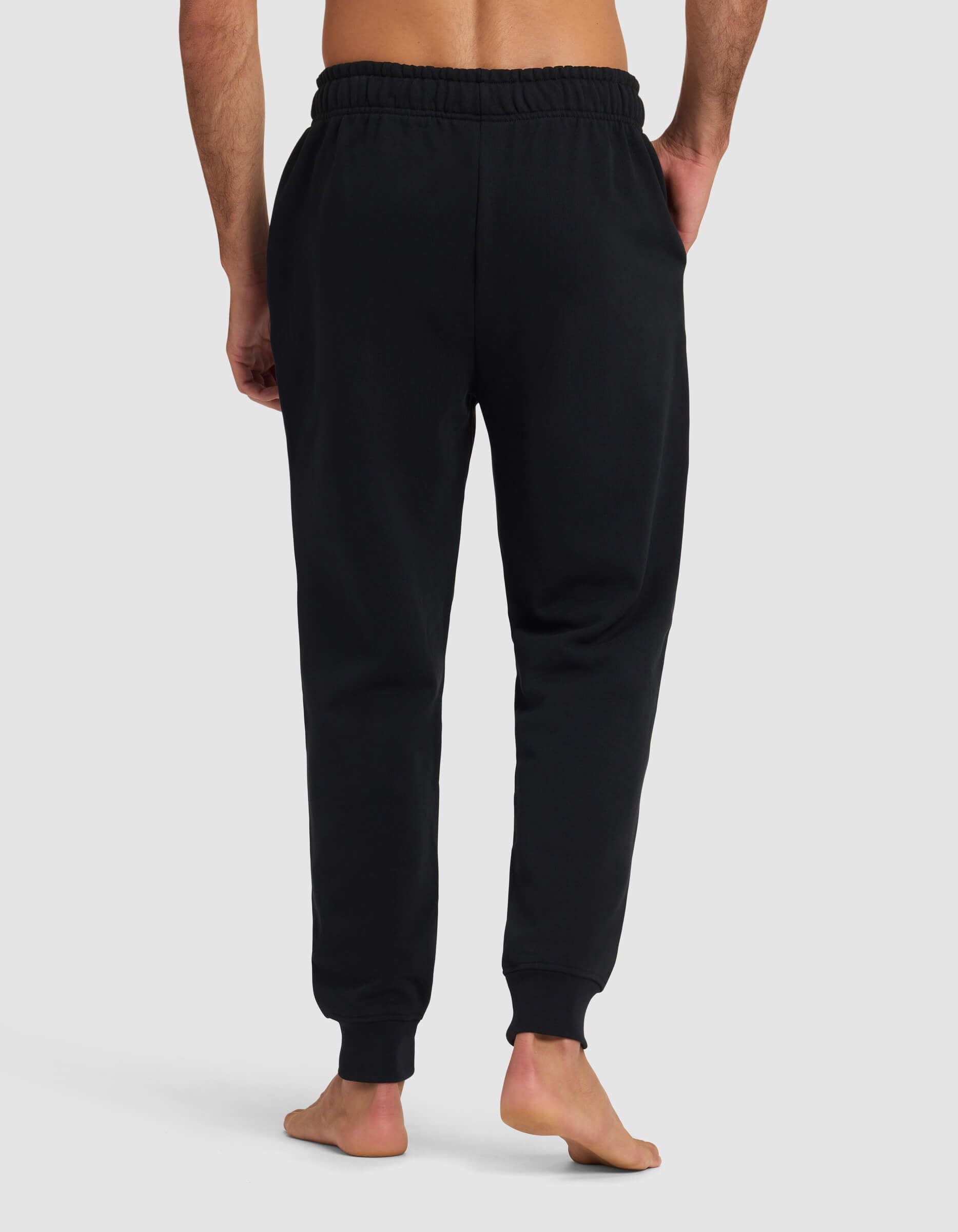 A barefoot man stands facing away, wearing Cozy Earth Men's Dylan Jogger—black sweatpants with an elastic waistband and cuffs—against a plain light gray background.