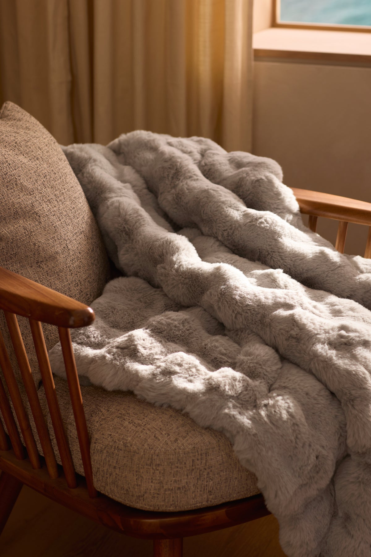 The Cozy Earth Bubble Cuddle Blanket, in plush light gray faux fur, is draped over a cushioned wooden chair by a window with beige curtains, adding warmth and inviting comfort to the room. |Color:Light Grey