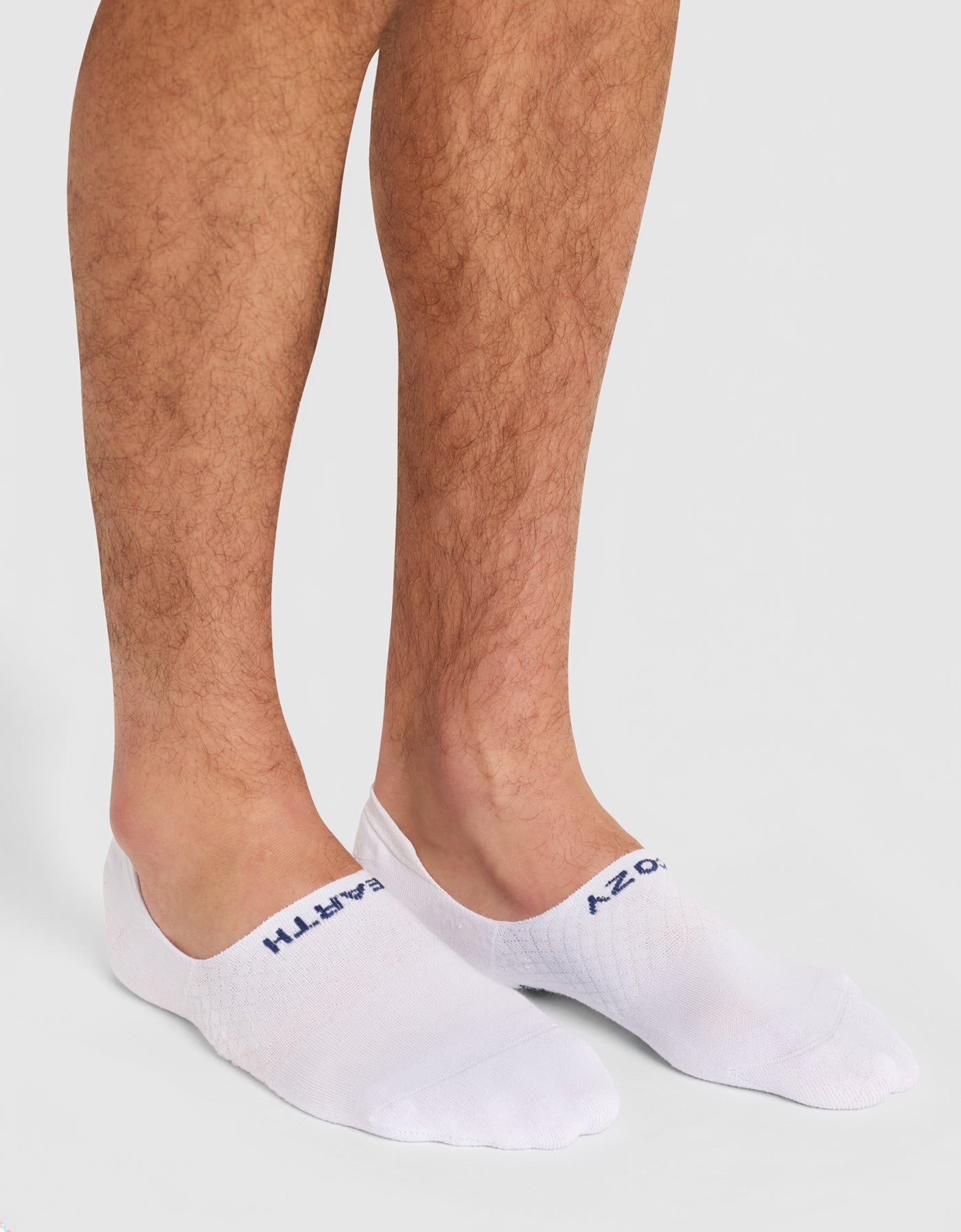A person wears Cozy Earth Men's Essential No-Show Socks (white with blue text) while standing on a light gray surface; only their lower legs and feet are visible.|Color:White