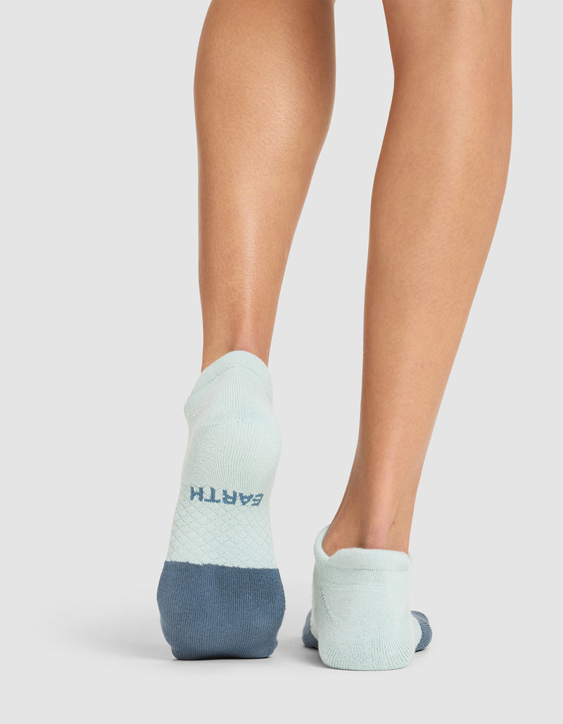 A person is shown from behind wearing Cozy Earth's Essential Ankle Socks in light and dark blue, with "EARTH" printed on the heel of the left sock, standing on a light-colored surface.