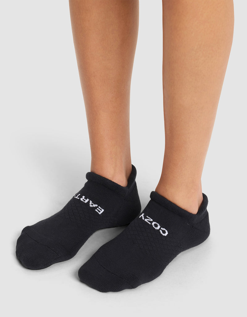 A person wearing Cozy Earth's Essential Ankle Socks stands on a light gray surface. The black socks feature white "COZY" text on the right foot and "EARTH" on the left.