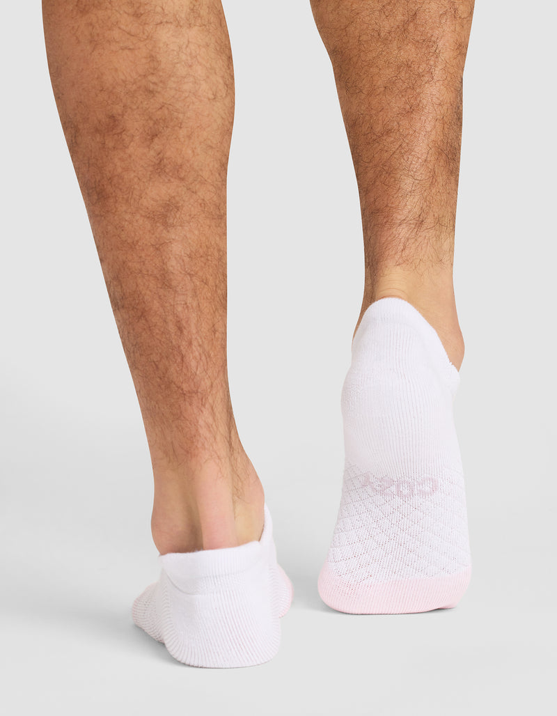A person with light brown, hairy legs is shown from behind, wearing Cozy Earth's Essential Ankle Sock in white with pink accents, standing on a light gray surface.