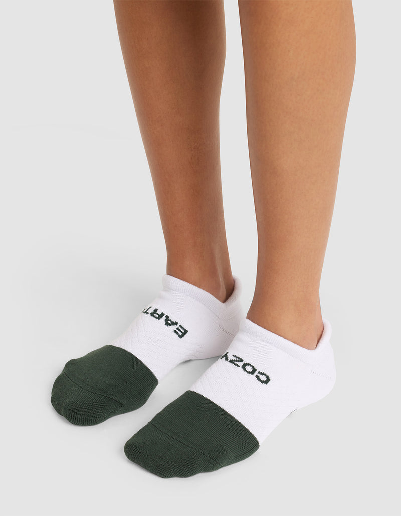 Someone stands on a light surface wearing Cozy Earth Essential Ankle Socks—white with dark green toes and "COZY EARTH" in black lettering on top.