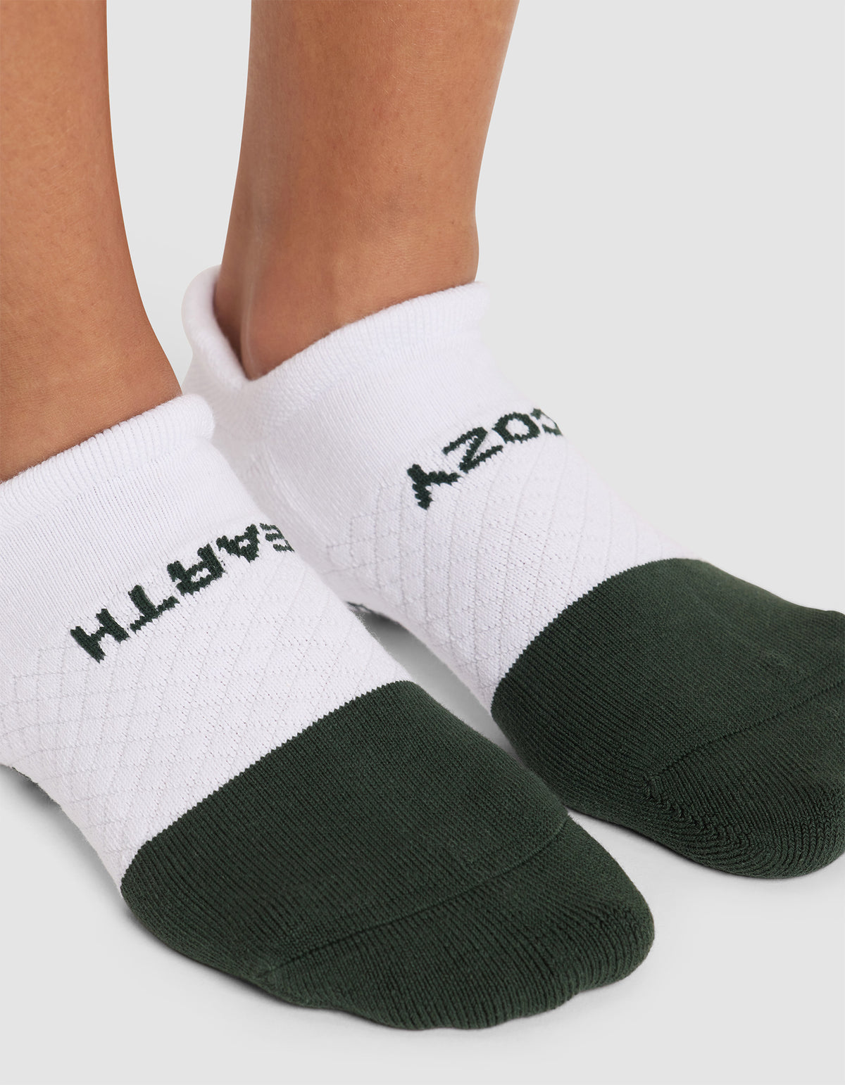 Close-up of feet wearing Cozy Earth's Essential Ankle Sock in white and dark green, featuring a textured pattern and partial black text reading “EARTH” and “COZY” on each sock. 