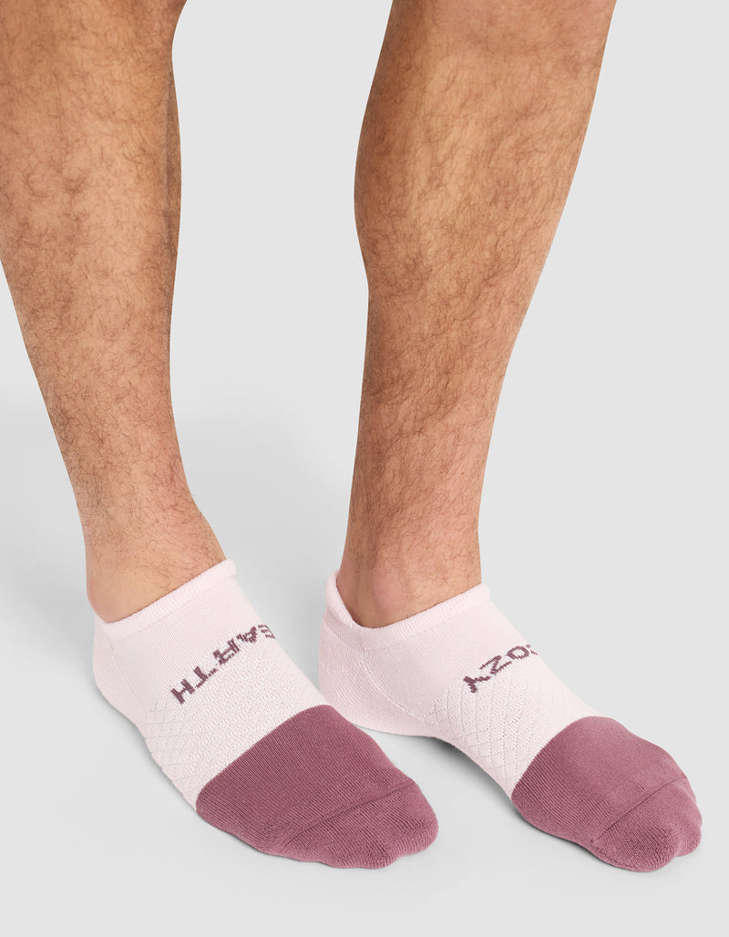 A person wearing Cozy Earth's Essential Ankle Socks in pink and purple, featuring "EARTHY COZY" on top, stands on a light gray surface.