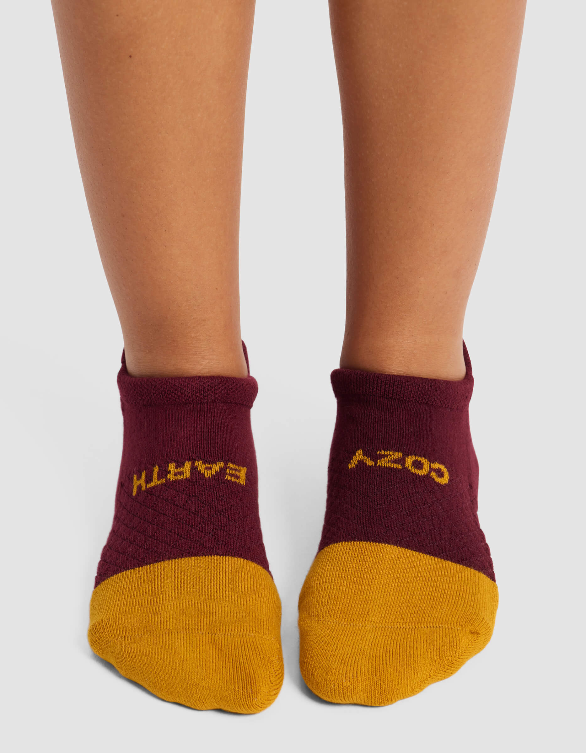 A person wears Cozy Earth’s Women's Essential Ankle Socks—maroon with yellow toes and upside-down "COZY" and "EARTH" text—standing on a light surface.|Color:Burgundy