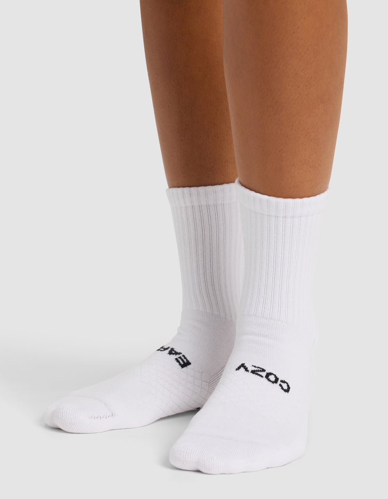 Someone wears Cozy Earth's Essential Calf Sock (4-Pack) in white, featuring ribbed crew design and black text on the toes. Only their lower legs and feet are visible on a white surface; the socks look clean and snug.