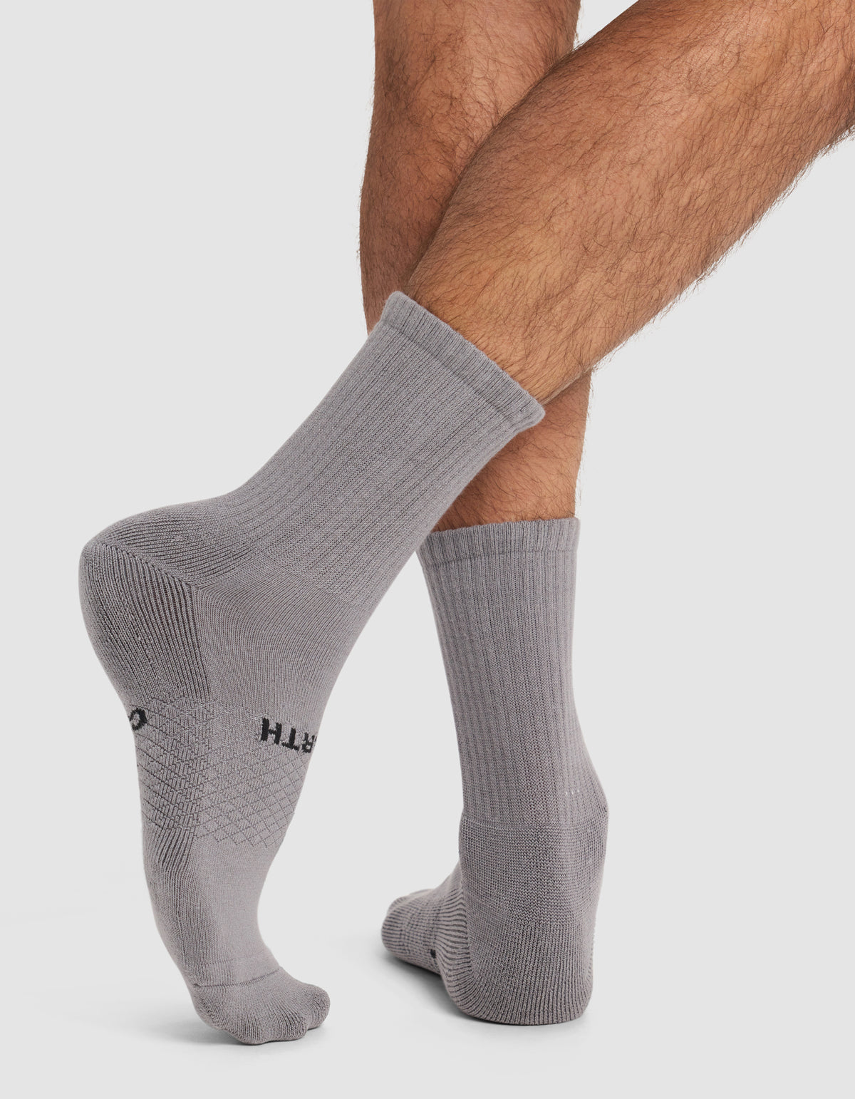 A person with crossed legs is wearing Cozy Earth’s Men’s Essential Calf Socks in light gray. The ribbed crew socks, featuring printed text on the soles, are displayed against a plain white background.