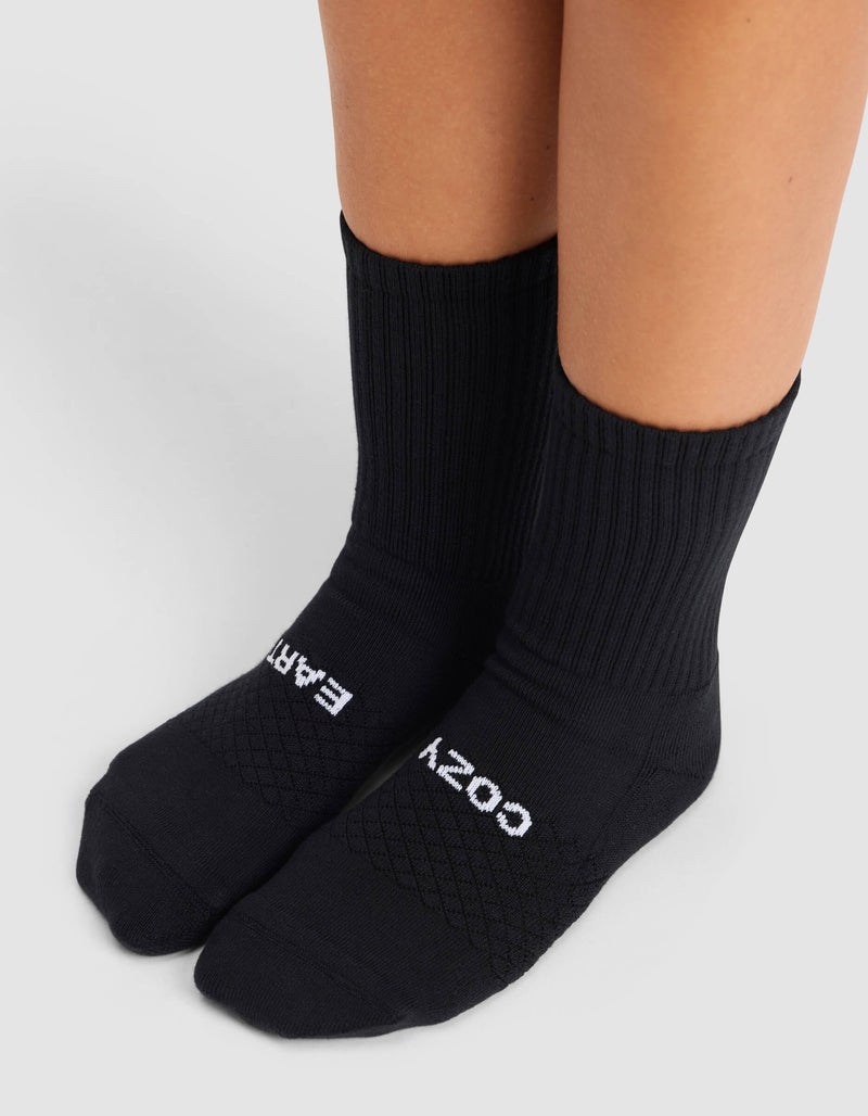 A person wearing Cozy Earth's Women's Essential Calf Sock in black, featuring "COZY EARTH" text on the tops of the feet, stands on a light gray surface.