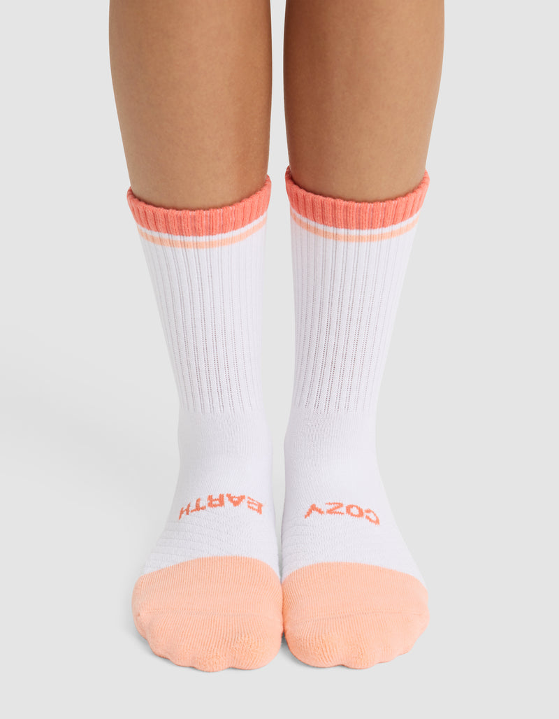 A person wearing Cozy Earth Essential Calf Socks in white with peach toes, heels, and cuffs. "COZY" is printed on the right sock and "BATH" on the left sock in peach lettering near the toes.