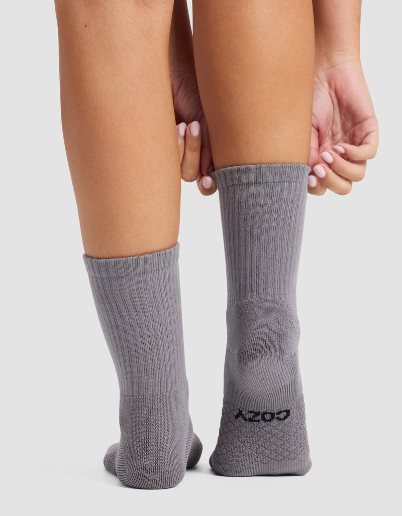 A woman with manicured nails and light skin stands on a white surface, wearing gray Women's Essential Calf Socks by Cozy Earth. "Cozy Earth" is printed in black on the heel of one sock.